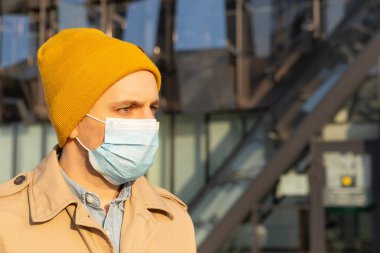 Yüzünde tıbbi maske olan, bahar kıyafetleri içinde yakışıklı bir Avrupalı. Grip virüsü veya koronavirüs enfeksiyonuna karşı korunmak için solunum cihazındaki bir adamın portresi..