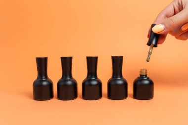 Bej arka planda siyah kavanozlardaki ojeler. Kadın eli, oje sürmek için fırça tutuyor..