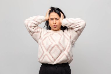 Kışlık kazak giymiş güzel bir esmerin portresi. Genç bayanın başı ağrıyor, gri arka planda. Kız ellerini başının arkasında tutuyor..