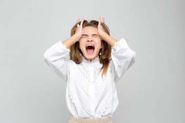 Başı ağrıyan genç bir kadın başını beyaz gri bir arka planda tutuyor. Kadın çok acı çekiyor.