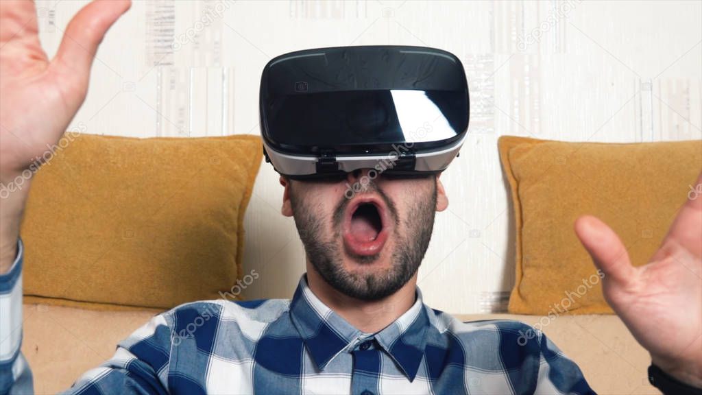 Bearded cheerful young man uses his VR virtual reality glasses at home on the couch