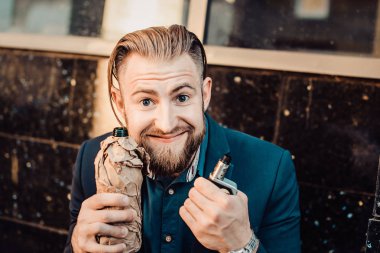 Takım elbiseli neşeli sakallı adam sarılmış şişe ve vape tutarak 