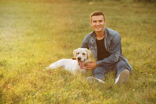 Porträt eines lächelnden Typen und seines Labradors, der bei Sonnenuntergang im Park auf Gras sitzt — Stockfoto