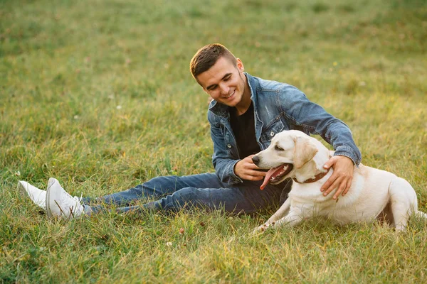 Улыбающийся мужчина сидит на траве со своим собачьим лабрадором в парке — стоковое фото