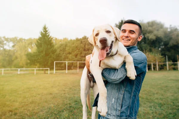 Porträt eines glücklichen Mannes, der seinen befreundeten Hund Labrador bei Sonnenuntergang im Park hält — Stockfoto