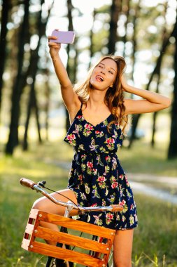 Bir bisiklet bir sepet ile güzel kız ormanda bir akıllı telefon üzerinde selfie yapar.