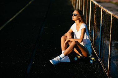 Güzel bir kız çitin onun kaykay üzerinde günbatımı üzerinde oturan güneş gözlüğü.