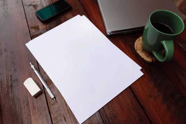 Lugar de trabajo, hoja de papel en blanco con un bolígrafo, taza de café y teléfono móvil y portátil en el escritorio de madera - Mock up — Foto de Stock