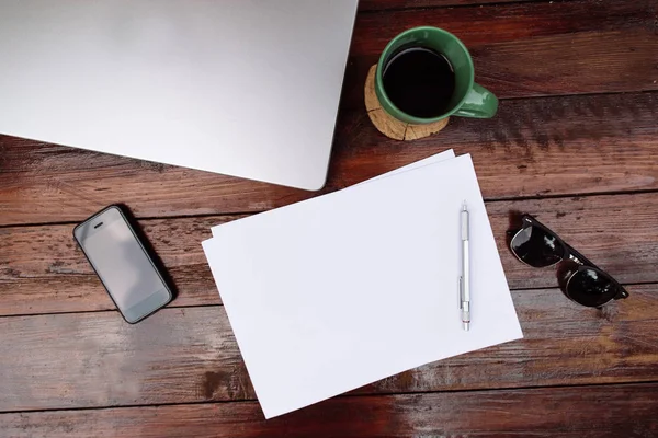 Lugar de trabajo, hoja de papel en blanco con un bolígrafo, taza de café y teléfono móvil y portátil en el escritorio de madera - Mock up — Foto de Stock