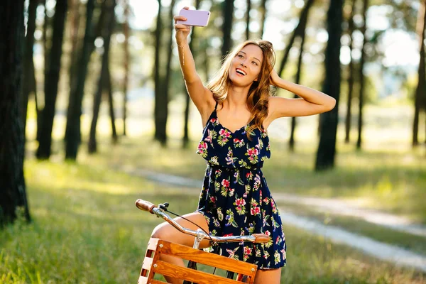 Hermosa chica en una bicicleta con una cesta hace selfie en un teléfono inteligente en el bosque . — Foto de Stock