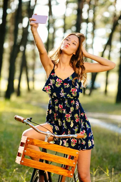 Hermosa chica en una bicicleta con una cesta hace selfie en un teléfono inteligente en el bosque . — Foto de Stock