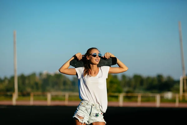 Hermosa mujer sostiene un monopatín en sus hombros con sus gafas de sol en . — Foto de Stock