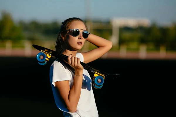 Een mooi meisje in sundlasses houdt een skateboard terwijl het controleren van haar haren. — Stockfoto