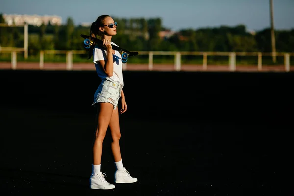 Una hermosa chica sostiene un monopatín en el hombro al atardecer . — Foto de Stock