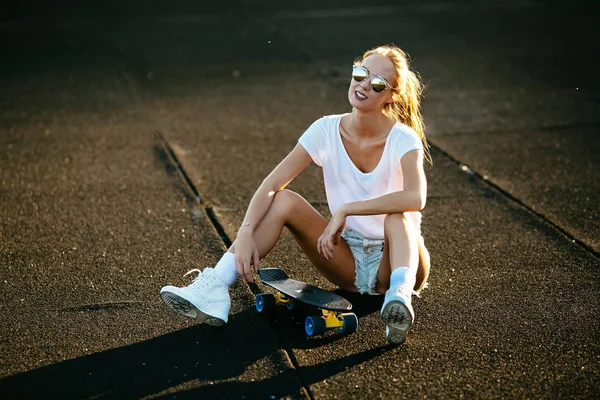 Genç kadın bir kaykay üzerinde onun güneş gözlüğü ile günbatımı sırasında oturur. — Stok fotoğraf