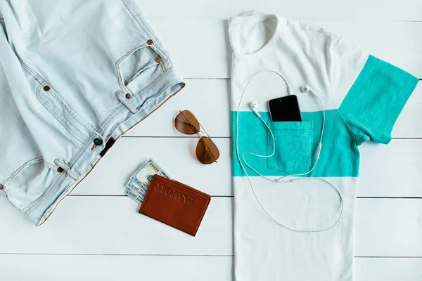Комплект мужской туристической летней одежды с паспортом, деньгами, солнцезащитными очками и смартфоном на белом деревянном фоне — стоковое фото