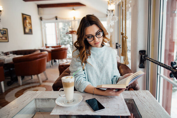 Женщина в очках читает книгу в кафе, когда пьет кофе
 