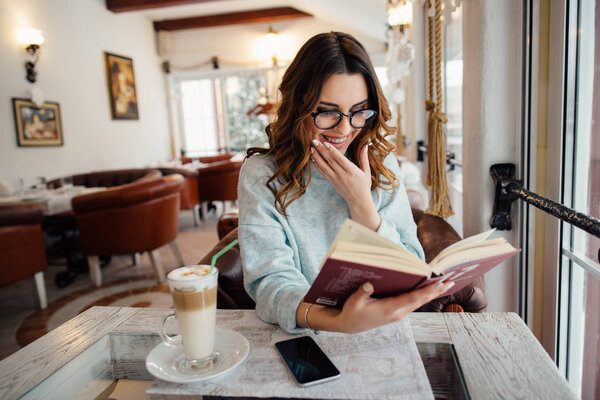 Улыбающаяся девушка в очках читает книгу в кафе
