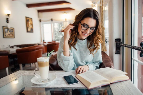 Glückliches junges Mädchen mit Brille liest interessantes lustiges Buch im Café — Stockfoto