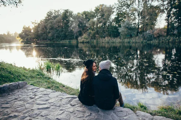 Mooie paar verliefd in warme kleren zit op riverside bij zonsondergang — Stockfoto