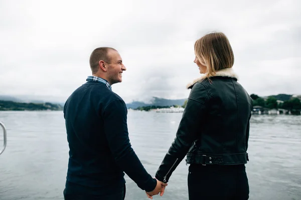 Jeune couple souriant se tenant la main et se regardant sur le front de mer avec une belle vue sur le lac et la montagne nuageuse — Photo
