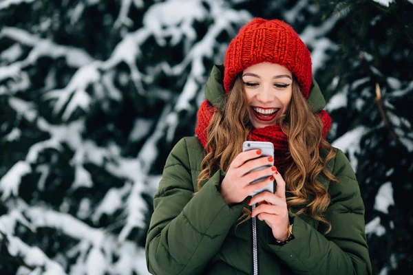 Chica en sombrero de invierno rojo y bufanda con teléfono inteligente y sonriente . — Foto de Stock