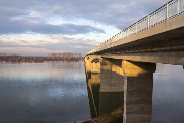 Kuvvetlendirilmiş beton köprü, büyük bir nehrin yanından geçen yol ve araba trafiği için kullanılır, güneşli bir öğleden sonra, kışın alacakaranlıkta, kırsal kesimde iki nehir kıyısını birbirine bağlar.