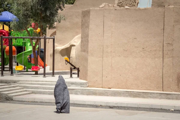 Shape of a woman walking in the streets of Yazd, Iran, passing near a children playground, wearing a niqab, an integral modest clothing veil covering the head and the body