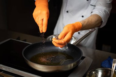 Ellerinde eldiven olan bir aşçı tavaya baharat koyar.
