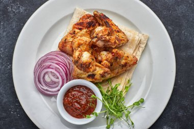 Izgara tavuk kanadı, barbekü sosu, pide ekmeği, mikroyeşil ve soğan halkaları, beyaz tabakta, koyu bir arka planda. Üst görünüm