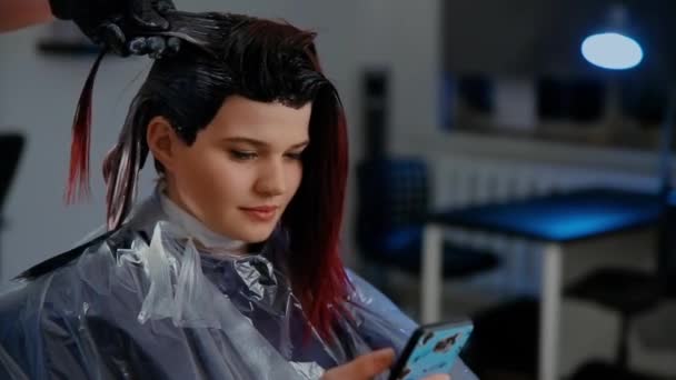 Concepto de belleza, peinado y gente - mujer joven feliz con teléfono inteligente y peluquería haciendo peinado en el salón — Vídeo de stock