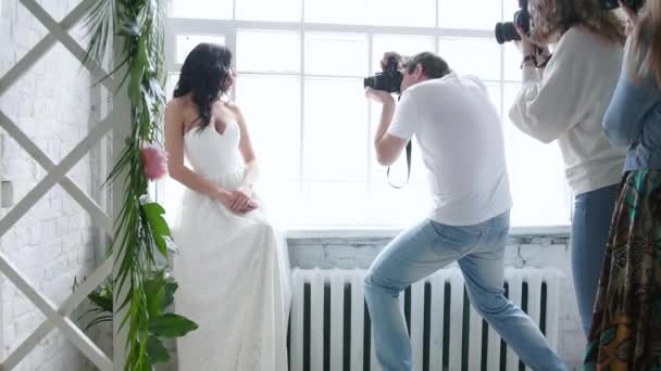 Φωτογράφοι που φωτογραφίζει μια όμορφη μελαχρινή νύφη μοντέλο. Σε ένα νυφικό σε ένα master class. Η κοπέλα ποζάρει δίπλα στο παράθυρο. Φωτογραφίες εκπαιδεύονται. — Αρχείο Βίντεο
