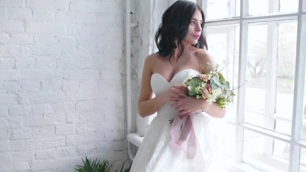 Sposa bella bruna in piedi in abito da sposa con bouquet vicino alla finestra — Video Stock