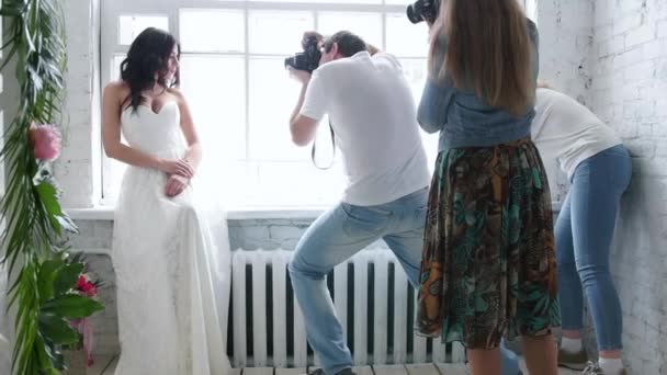 Fotógrafos fotografando uma linda modelo de noiva morena. Em um vestido de noiva em um master class. A menina está posando pela janela.Fotografias são treinadas . — Vídeo de Stock