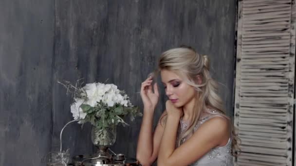 Belle femme blonde modèle avec une robe de mariée s'asseoir sur une chaise avec un bouquet de fleurs sur un fond gris . — Video