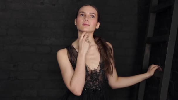 Beautiful brunette in a black dress posing and looking at the camera on a black wall background. — Stock Video