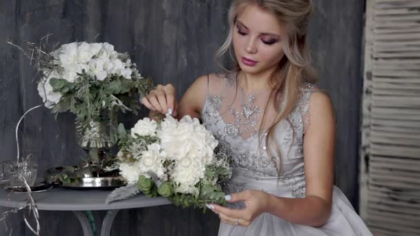 Belle femme blonde modèle avec une robe de mariée s'asseoir sur une chaise avec un bouquet de fleurs sur un fond gris — Video