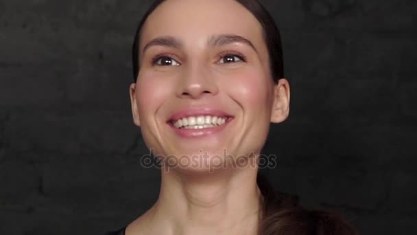 A cute girl in a black dress against a brick wall background laughs and looks at the camera. — Stock Video