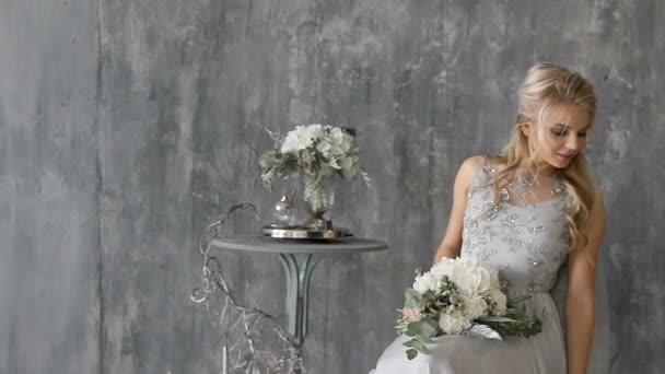 Belle femme blonde modèle avec une robe de mariée s'asseoir sur une chaise avec un bouquet de fleurs sur un fond gris — Video