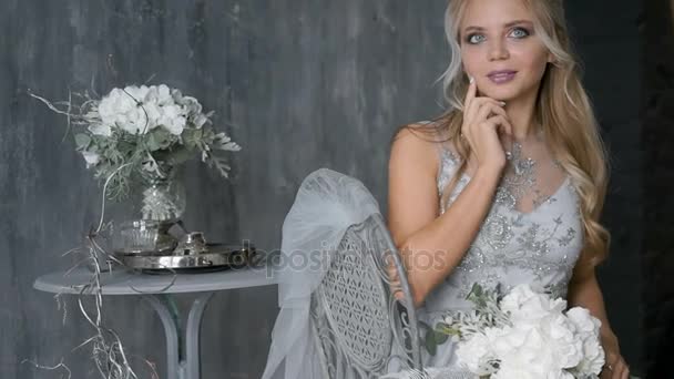 Belle femme blonde modèle avec une robe de mariée s'asseoir sur une chaise avec un bouquet de fleurs sur un fond gris — Video