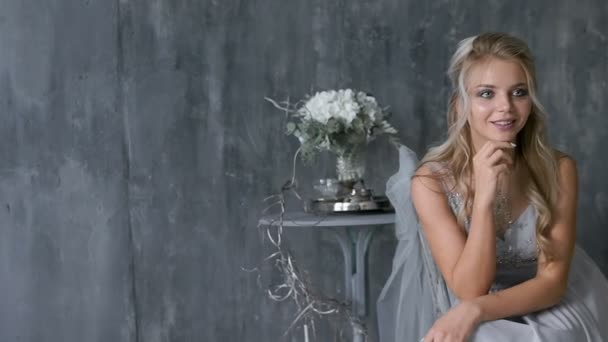 Красива блондинка модель з весільною сукнею сидить на стільці з букетом квітів на сірому фоні — стокове відео