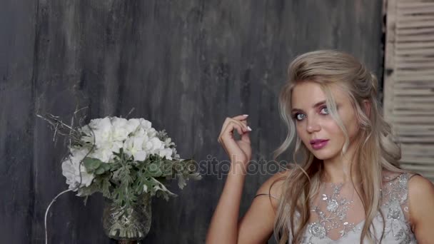Красивая блондинка-модель в свадебном платье сидит на стуле с букетом цветов на сером фоне . — стоковое видео