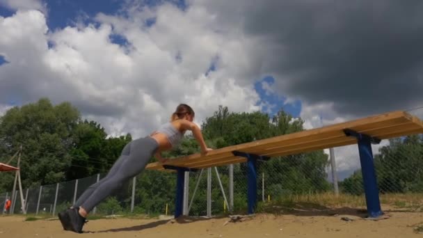 Vista de perfil de una atleta haciendo algunos descensos de tríceps en un banco del parque — Vídeos de Stock