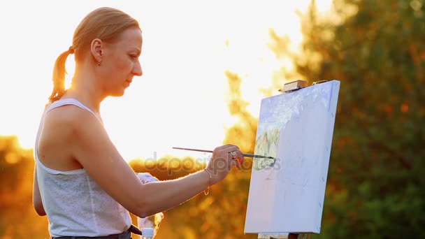 Ritratto di una ragazza con i capelli bianchi in una t-shirt bianca, raffigurante un paesaggio su tela al tramonto al sole con pittura ad olio e pennello . — Video Stock