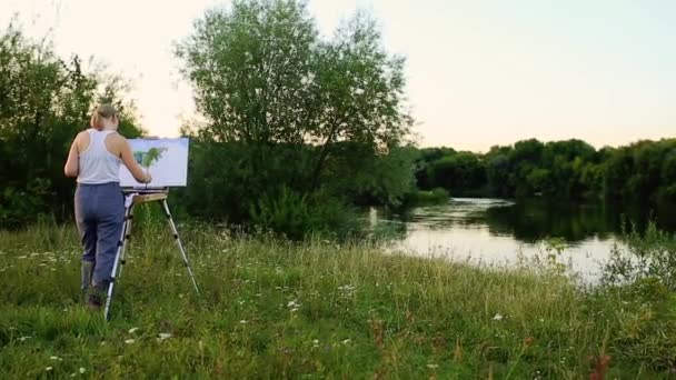 Una artista con un pincel y un polytray en sus manos dibuja sobre lienzo sobre el telón de fondo de la naturaleza. El artista se basa en el caballete. Artista en el trabajo — Vídeo de stock