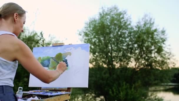 Молодая женщина с длинными волосами, которая развивается на ветру, рисует картину на холсте, который стоит на мольберте. Леди на открытом воздухе, она черпает из жизни — стоковое видео