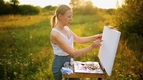 Hermosa mujer rubia artista con un pincel en la mano dibuja sobre lienzo en la naturaleza — Vídeos de Stock