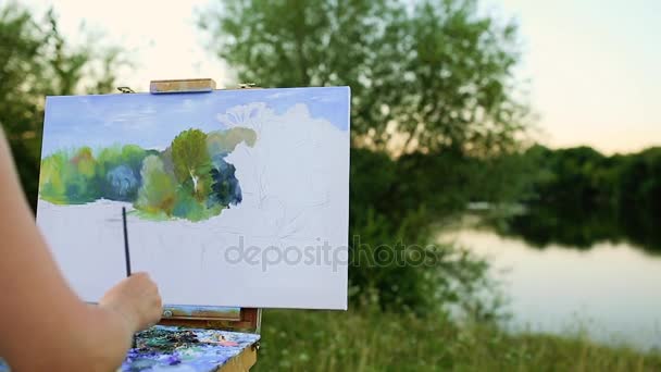Молодий художник малює картину в парку з пензлями і кольорами, сидячи біля романтичного озера і міських будівель на задньому плані. ультраширокий кут огляду — стокове відео