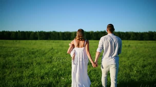 Glückliche Familie. Papa Mama und ein kleines Mädchen, die in einem Feld in weiß gekleidet unter den Strahlen der untergehenden Sonne gehen — Stockvideo