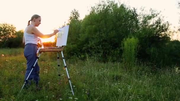 A woman artist with a brush and polytray in her hands draws on canvas against the backdrop of nature. The artist draws on the easel. Artist at work — Stock Video